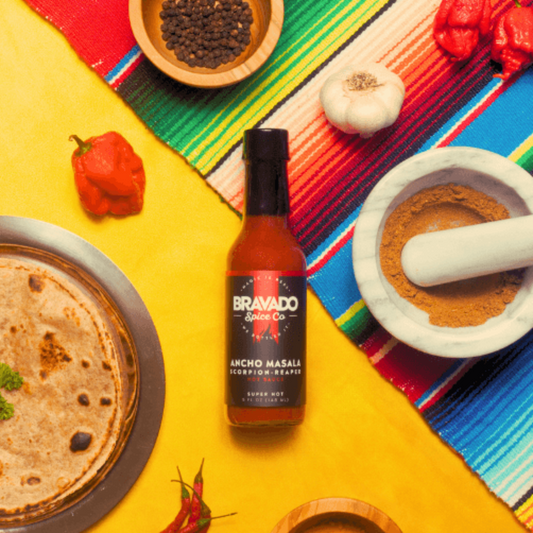 Ancho Masala Scorpion Reaper hot sauce bottle on colorful tablecloth and yellow background with reaper peppers, garlic, and tortillas.