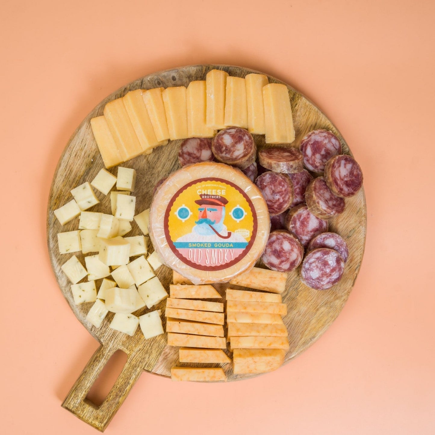 Wooden cheese board with various cheeses and sliced sausage on peach background