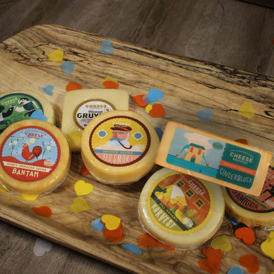 An assortment of cheese blocks such as 'Honey Sriracha Gouda' and 'Cinderblock Smoked Cheddar', presented on a wood cutting board surrounded by decorative elements of paper cutout hearts