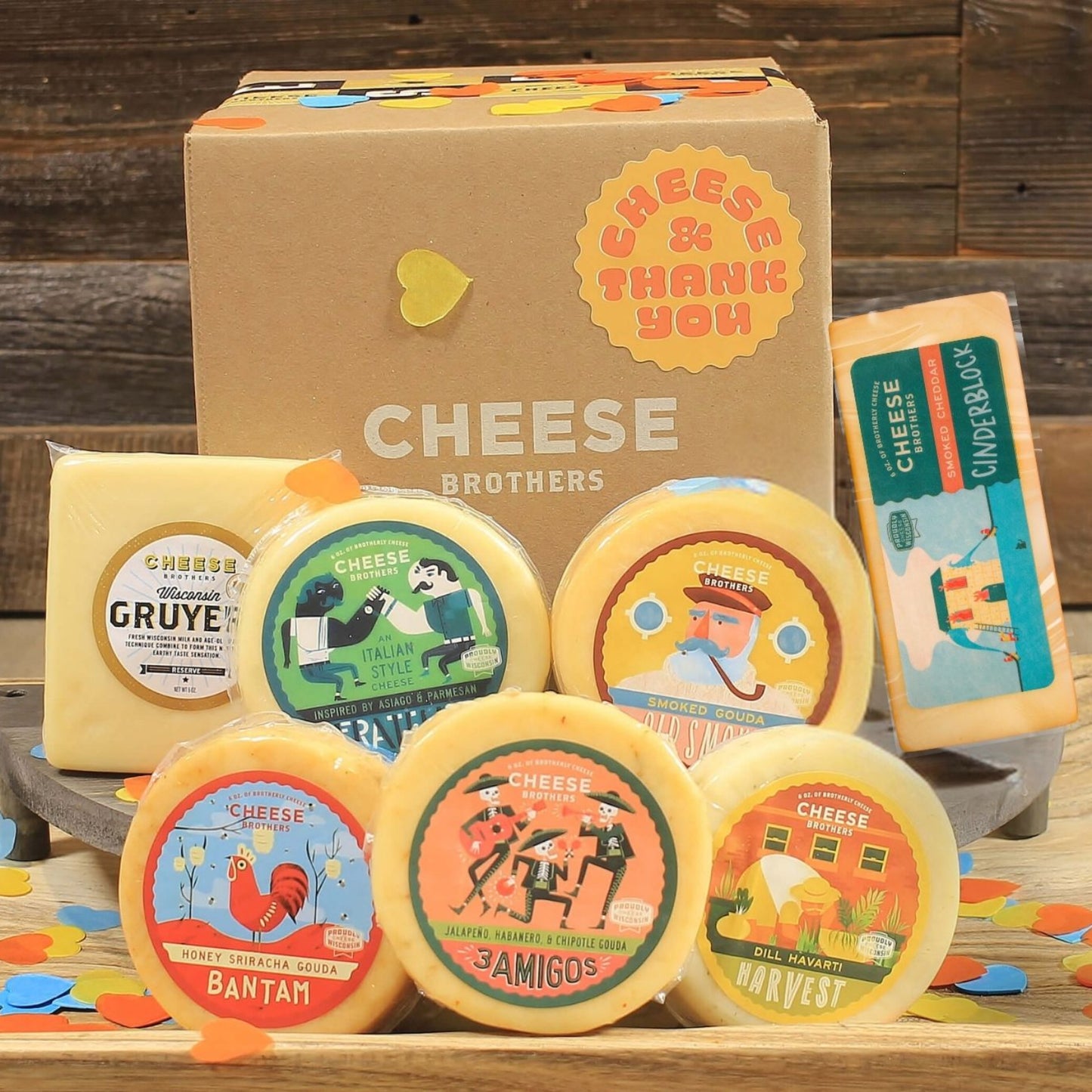 An assortment of cheese blocks presented on a wooden surface including various flavors like smoked gouda, dill havarti with decorative elements like a wooden serving platter, cut out paper hearts, and shipping box