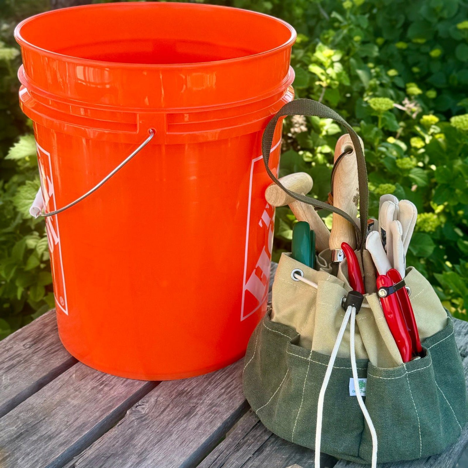 Orange bucket and green tool pouch with gardening tools on a wooden surface with greenery in the background