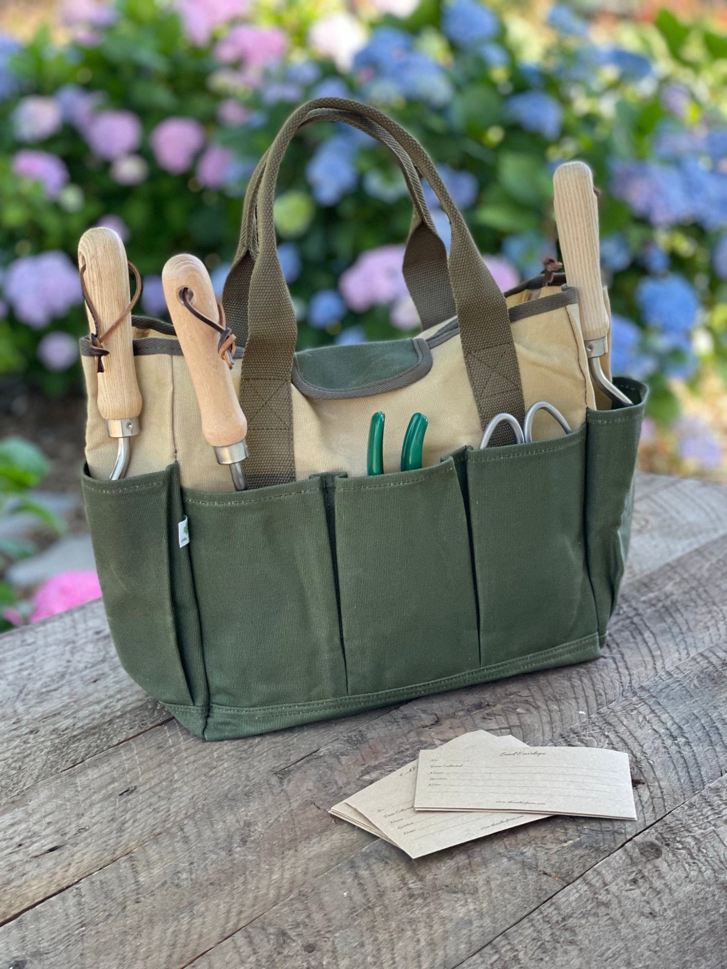 A green garden tool bag made of heavy waxed canvas, displayed on a wooden surface with gardening tools and paper gloves inside it, against a blurred floral background.