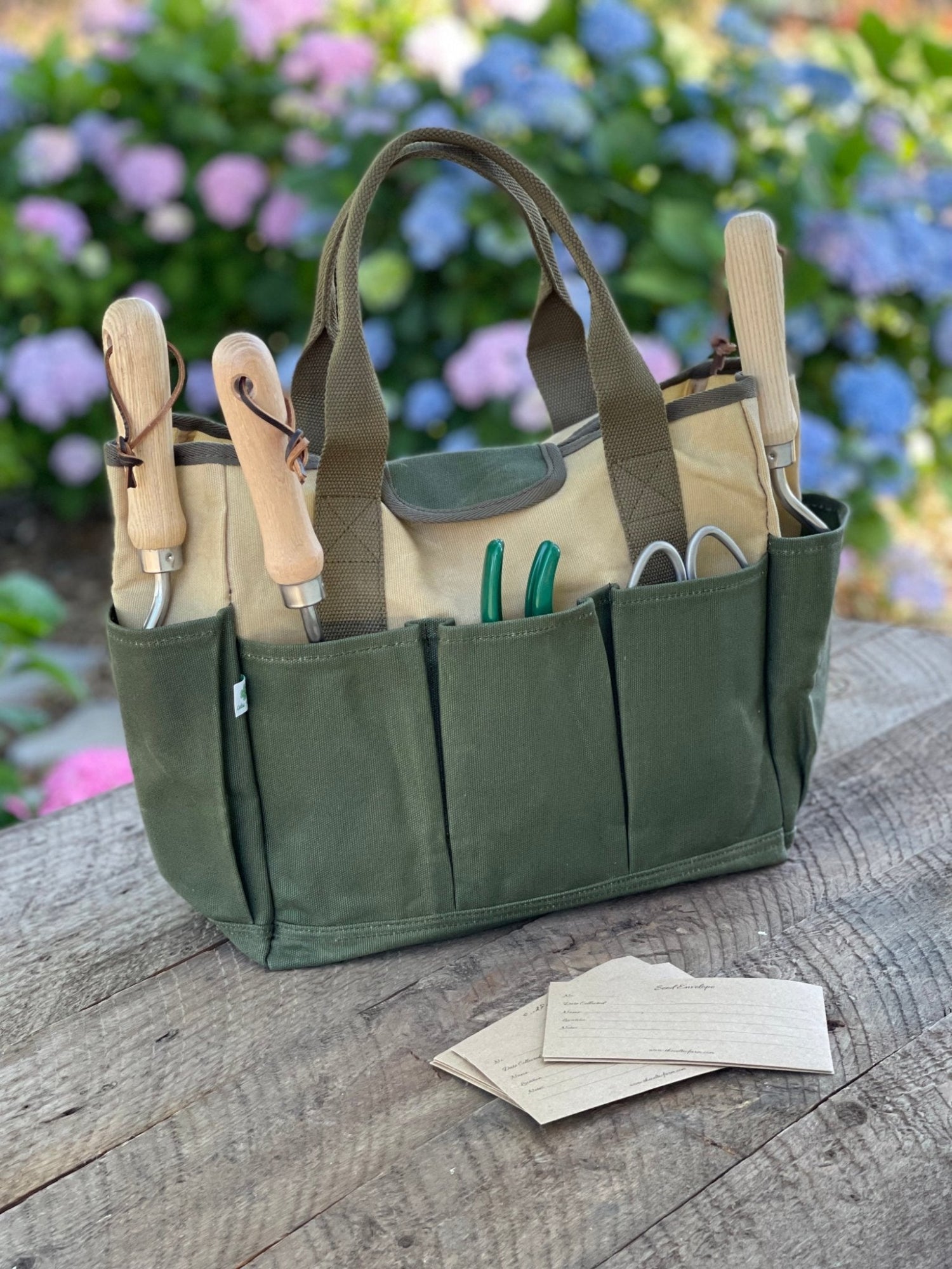 A green garden tool bag made of heavy waxed canvas, displayed on a wooden surface with gardening tools and paper gloves inside it, against a blurred floral background.