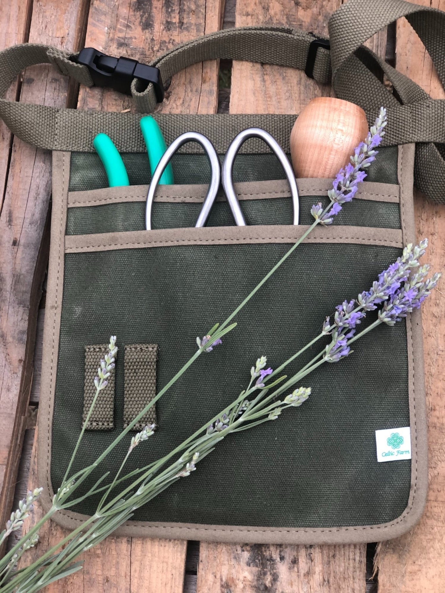 A green waxed canvas garden tool belt with two pockets and a cotton webbing belt, displayed with gardening tools and lavender sprigs.
