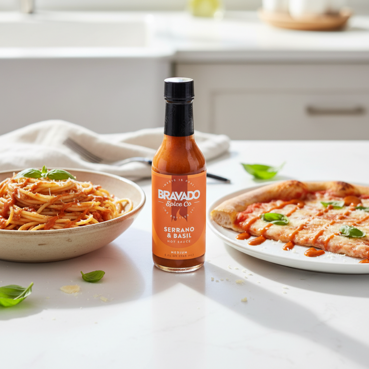 Serrano and Basil Hot Sauce bottle on a white kitchen counter beside a bowl of spaghetti and a plate of pizza drizzled with spicy blueberry hot sauce.