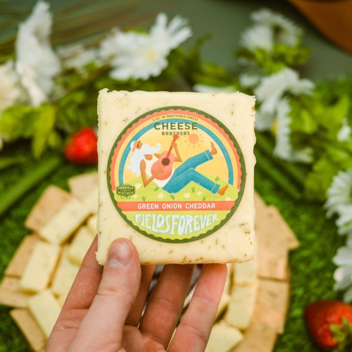 Hand presenting Green Onion Cheddar above a platter with crackers, sliced cheese, strawberries, flowers, and decorative grass
