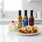 One bottle each of Pineapple Reaper, Blueberry Reaper, and Carolina Reaper hot sauces including in the Hottest Hot Sauce Gift Box on a white kitchen counter beside a plate of tacos and bowl of vanilla ice cream.