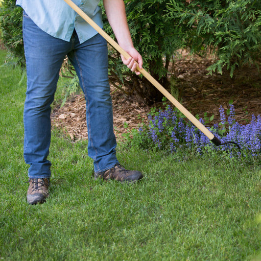 Person gardening with a Long Handle Weeder & Cultivator Garden Tool in a grassy area with plants and flowers.