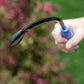 Hand holding a Mini Weeder & Cultivator Hand Tool  against a blurred green and pink outdoor landscape background