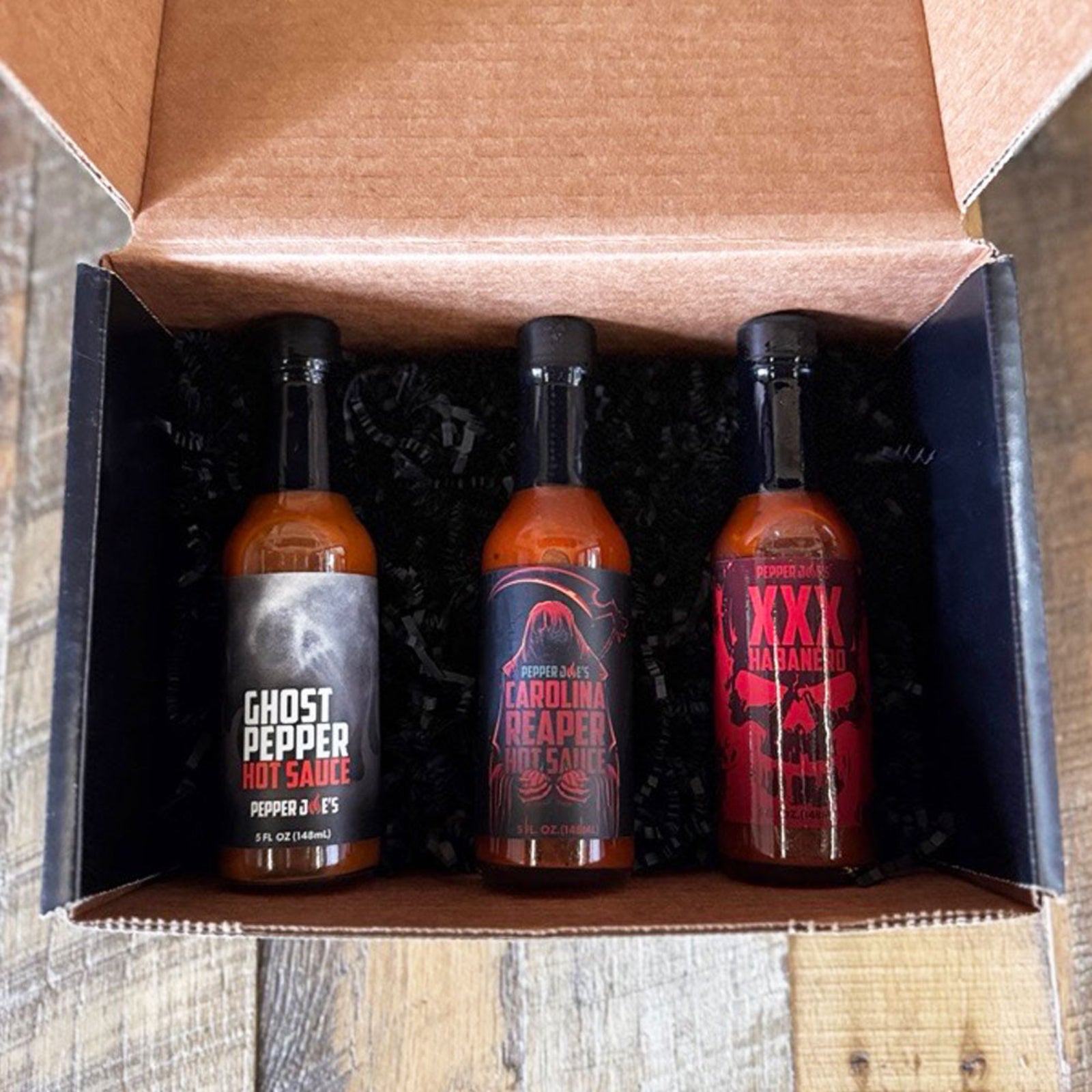 Pepper Joe's Tour of Heat hot sauce gift box that includes one bottle each of Ghost Pepper, XXX Habanero, and Carolina Reaper packaged inside gift box on a rustic background.