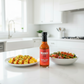 Árbol Chili & Garlic Hot Sauce bottle on a white kitchen counter beside a bowl of roasted vegetables and a plate of scrambled eggs drizzled with spicy hot sauce.