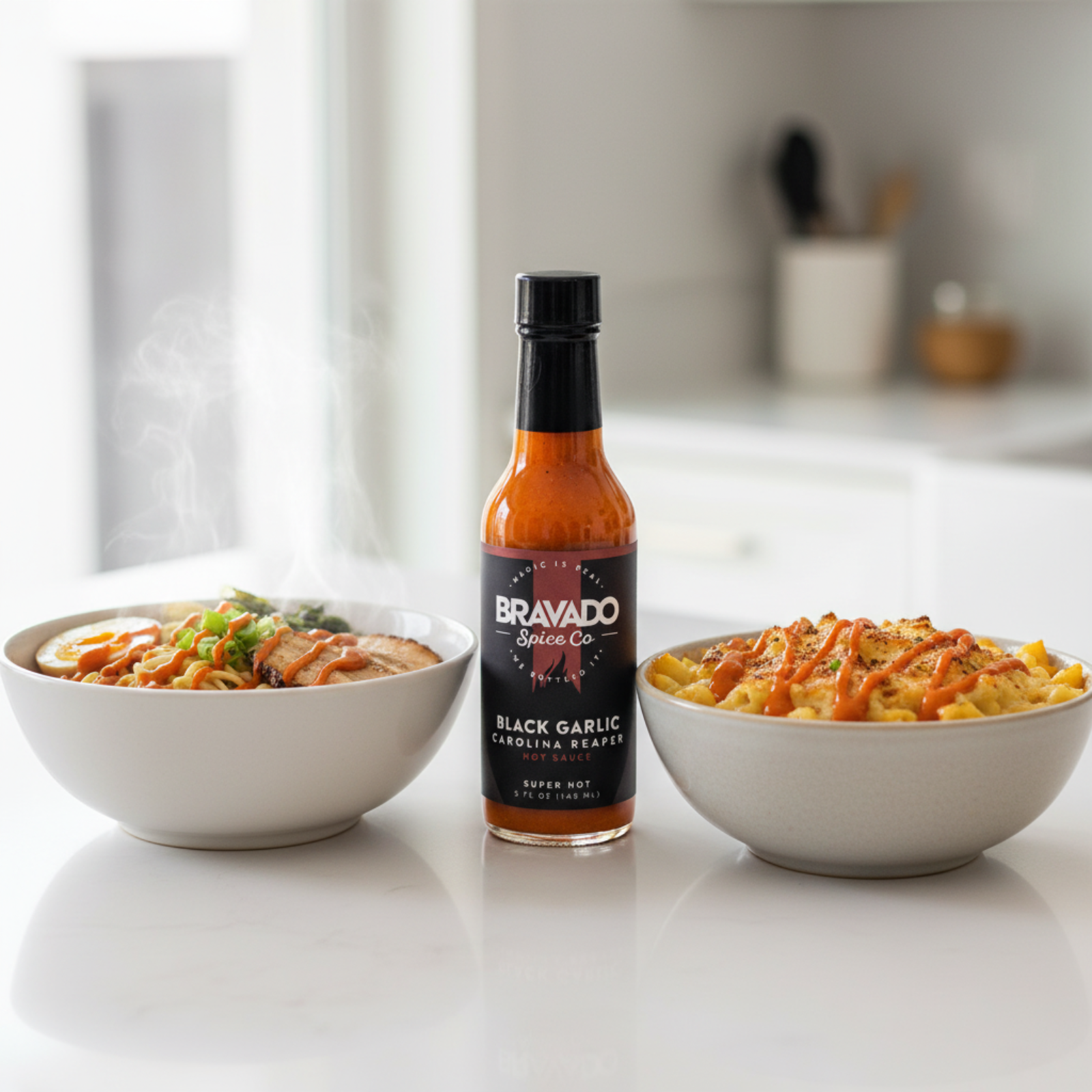 Black Garlic Carolina Reaper Hot Sauce bottle on a white kitchen counter beside a bowl of ramen and a bowl of mac and cheese drizzled with spicy hot sauce.