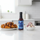 Blueberry Carolina Reaper Hot Sauce bottle on a white kitchen counter beside a bowl of ice cream and a plate of fried chicken wings drizzled with spicy blueberry hot sauce.