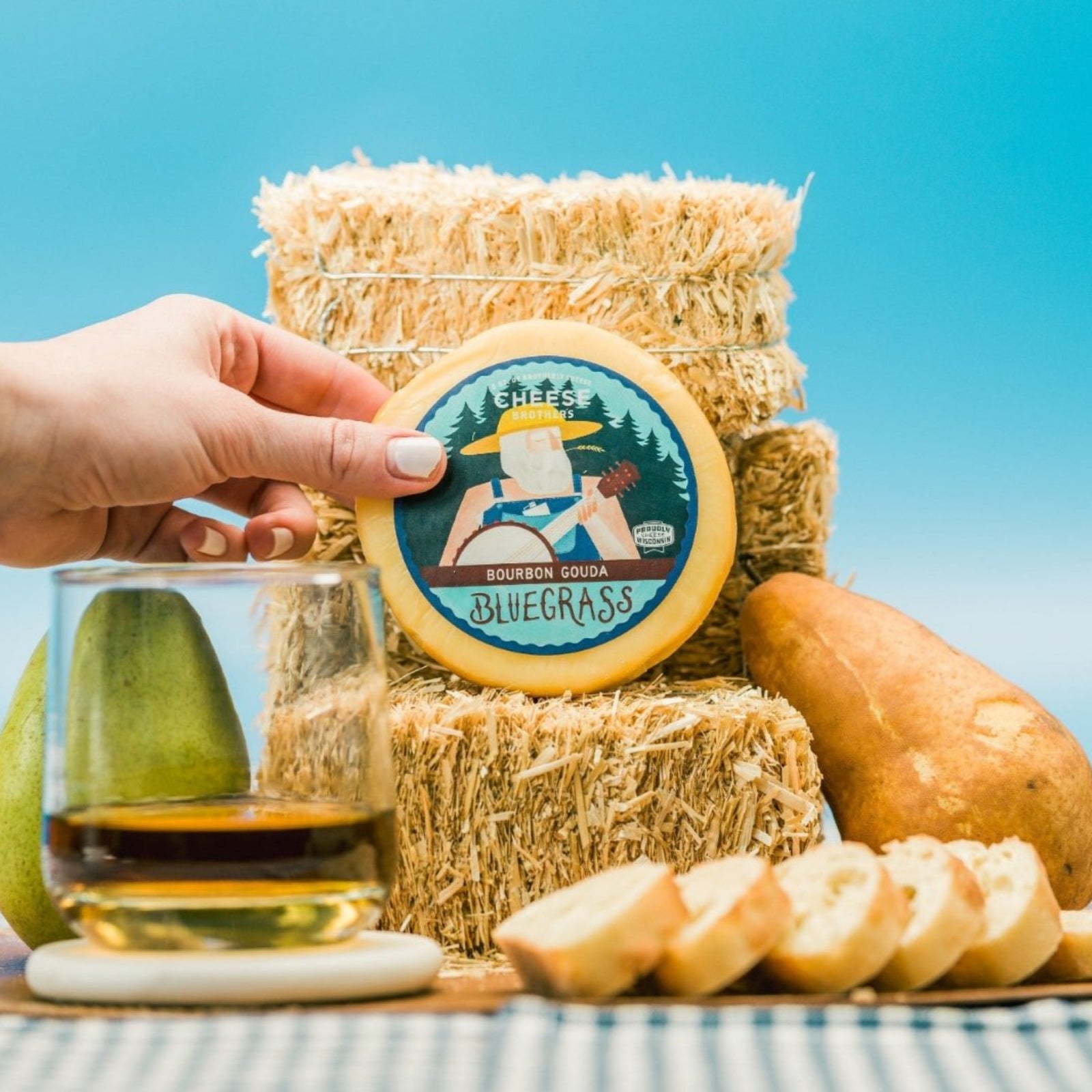 Bourbon Gouda block of cheese presented on small straw bales with decorative elements such as pears, sliced bread, and a glass of wine on a blue and white checkered tablecloth