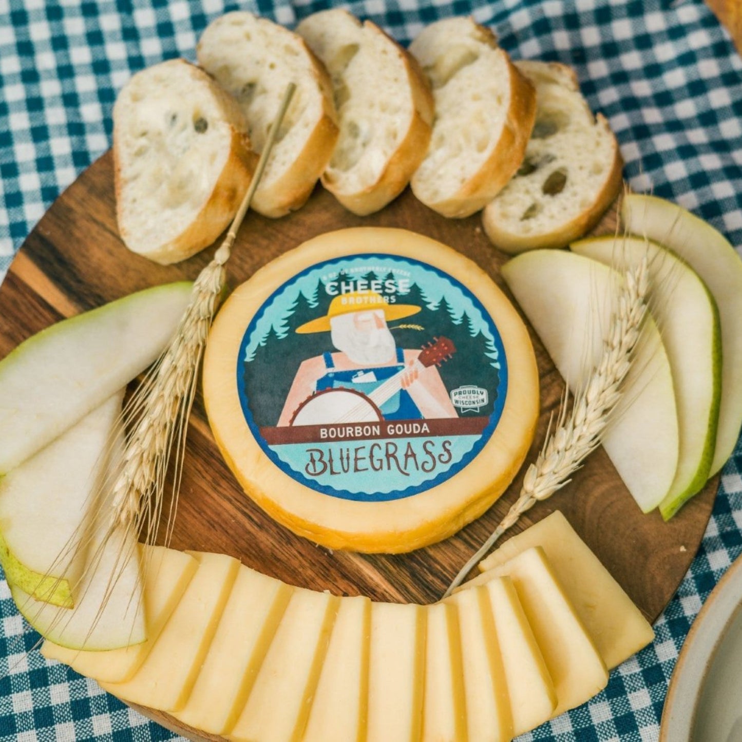 Bourbon Gouda block of cheese presented on a serving tray with decorative elements such as sliced pears, sliced cheese, and wheat on a blue and white checkered tablecloth
