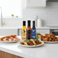 One bottle each of Pineapple Reaper, Carolina Reaper, and Blueberry Hot Sauce including in the Reaper Gift Box on a white kitchen counter beside a plate of pizza, plate of tacos, plate of fried chicken wings drizzled in the sauces.