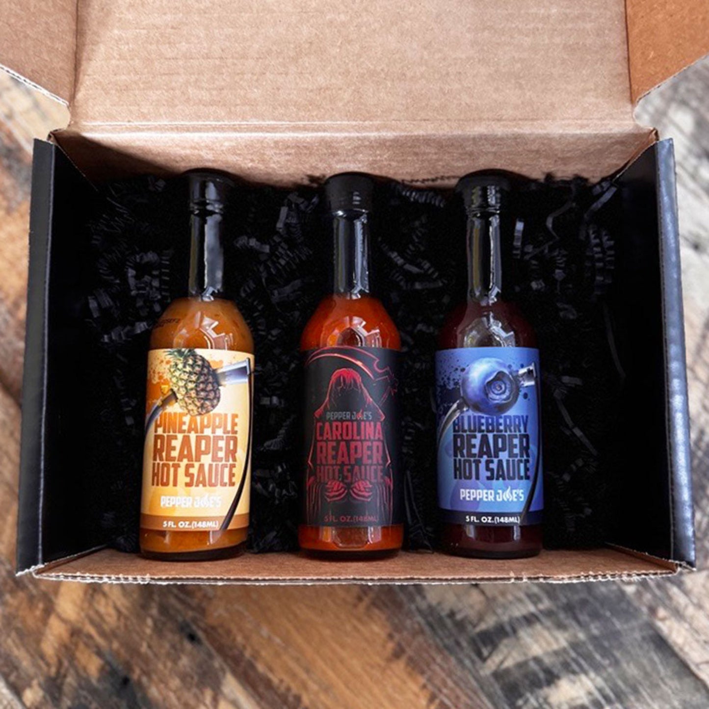 Pepper Joe's Carolina Reaper hot sauce gift box that includes one bottle each of Blueberry Reaper, Pineapple Reaper, and Carolina Reaper packaged inside gift box on a rustic background.