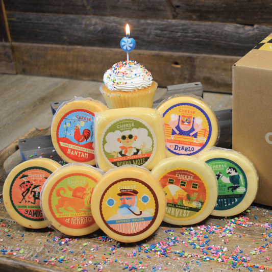 An assortment of cheese blocks presented next to a packed order in a box including various flavors like smoked gouda, fratello, dill havarti with decorative elements like a birthday cupcake and sprinkles