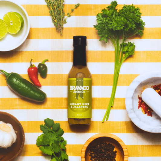 Bottle of creamy herb jalapeno hot sauce on a yellow and white striped tablecloth with herbs, jalapenos, and lime.