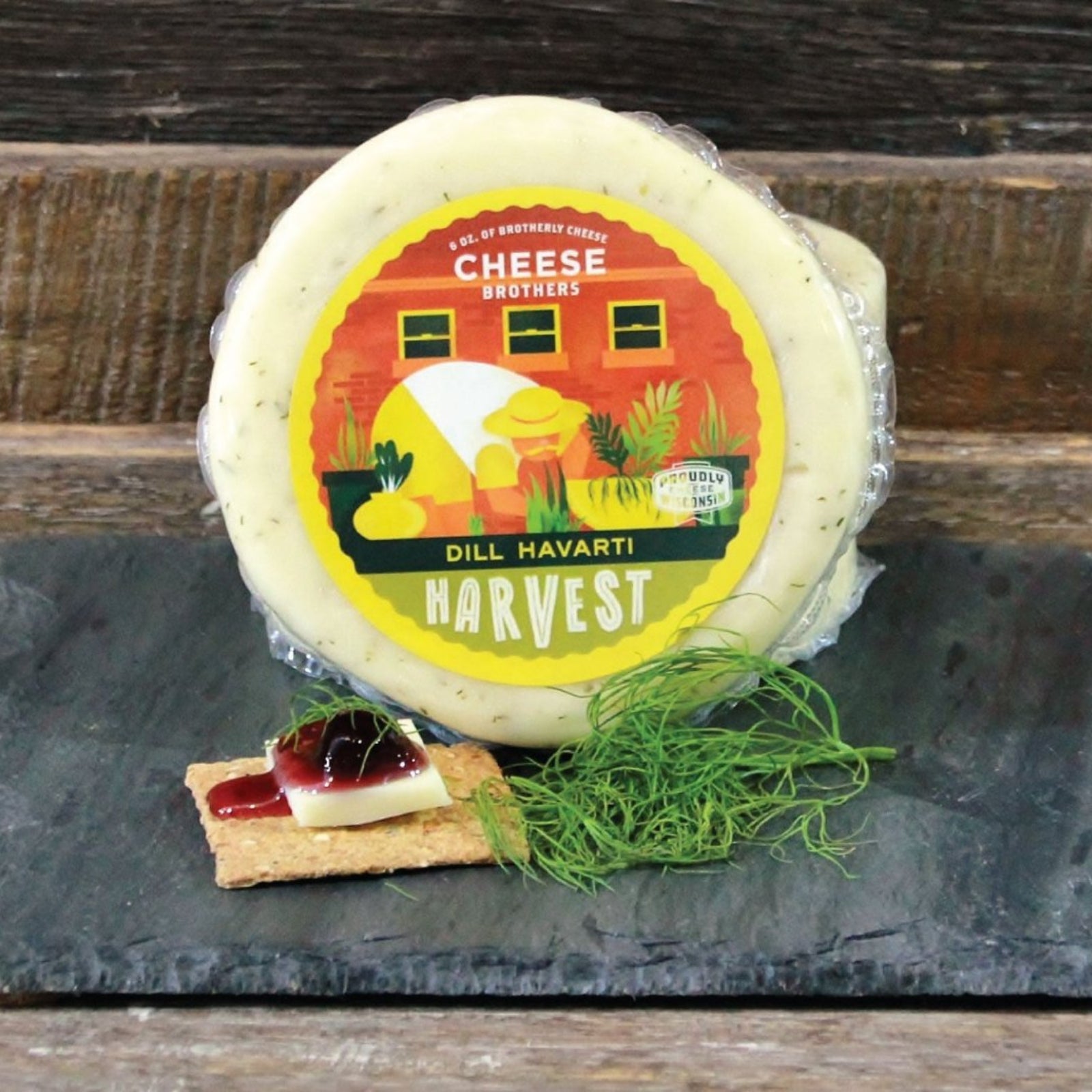 A wheel of Dill Havarti cheese on a slate platter presented with a small amount of dill herb  and a slice of cheese on a cracker topped with jelly.
