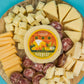 Wooden cheese board with dill havarti cheese wheel in the center and various types of cheese and cured meats sliced on a blue background.