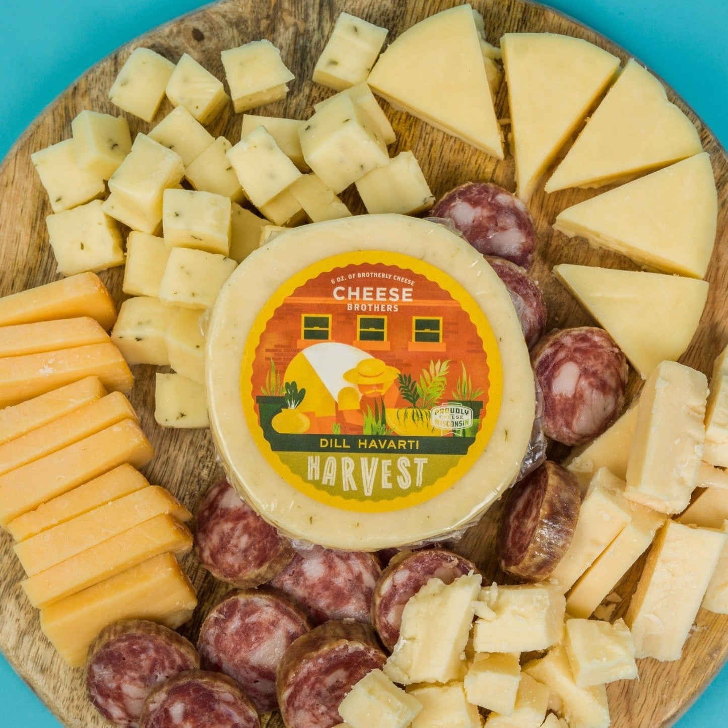 Wooden cheese board with dill havarti cheese wheel in the center and various types of cheese and cured meats sliced on a blue background.