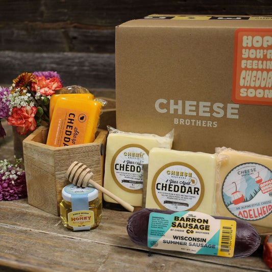 Feel cheddar soon gift box containing various types of cheese, including cheddar and smoked cheddar, with a honey dipper, all arranged in front of a cardboard box 