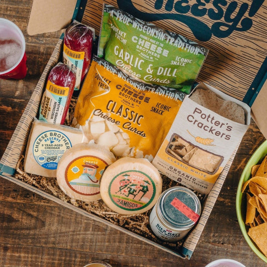 An assortment of gourmet snacks in a gift pack, including various cheeses, cheese curds, summer sausages, mustard, and crackers, displayed in an open gift box.