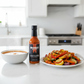 Garlic & Árbol Moruga Scorpion Hot Sauce bottle on a white kitchen counter beside a bowl of soup and a plate of grilled vegetables drizzled with hot sauce. 