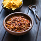 Bowl of chili with beans and ground meat on a wooden table, accompanied by tortilla chips with ghost pepper naga jolokia salt added to the chili.