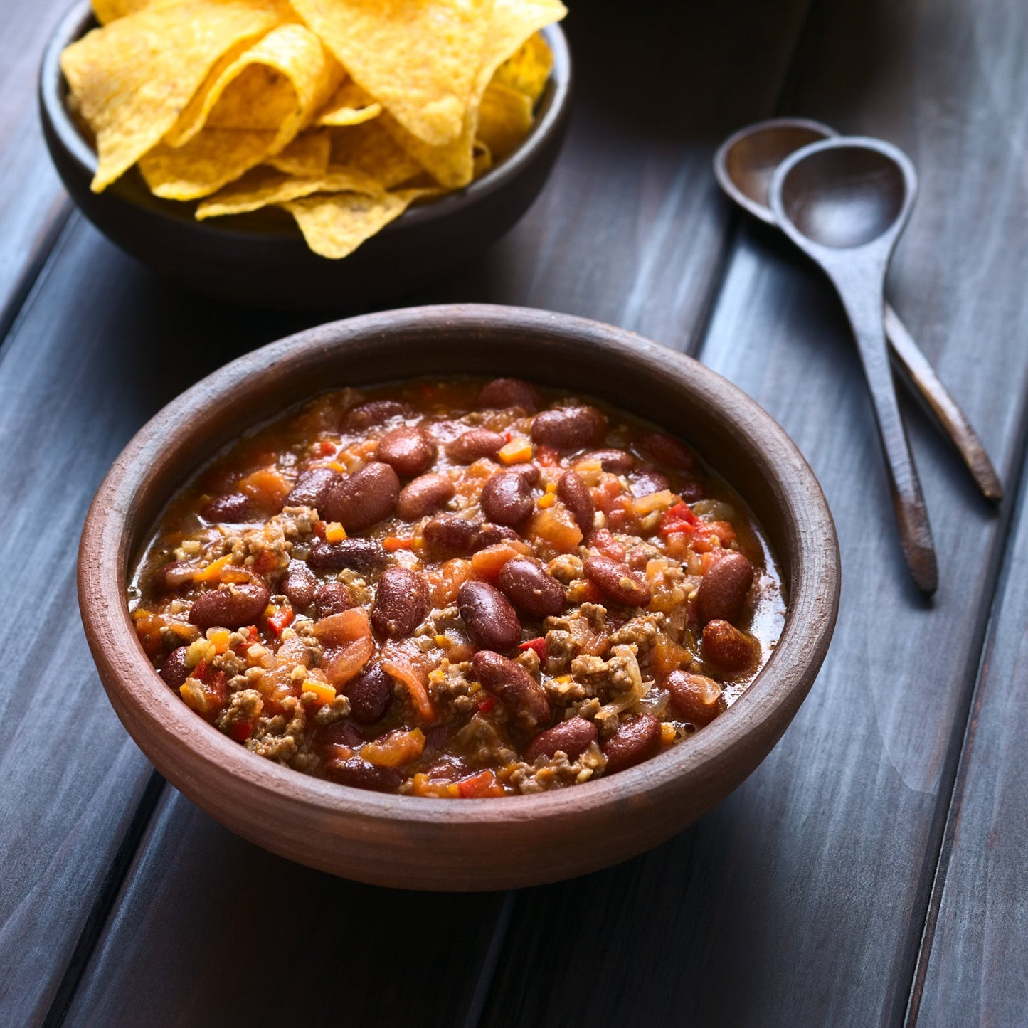 Bowl of chili with beans and ground meat on a wooden table, accompanied by tortilla chips with ghost pepper naga jolokia salt added to the chili.
