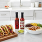 One bottle each of XXX Habanero and Garlic Habanero included in the Habanero Gift Box on a white kitchen counter beside a plate of tacos and rice bowl.
