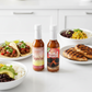 One bottle each of Tropical Habanero and Mango Habanero included in the Habanero Gift Box on a white kitchen counter beside a plate of tacos, two rice bowls, and a plate of grilled chicken.
