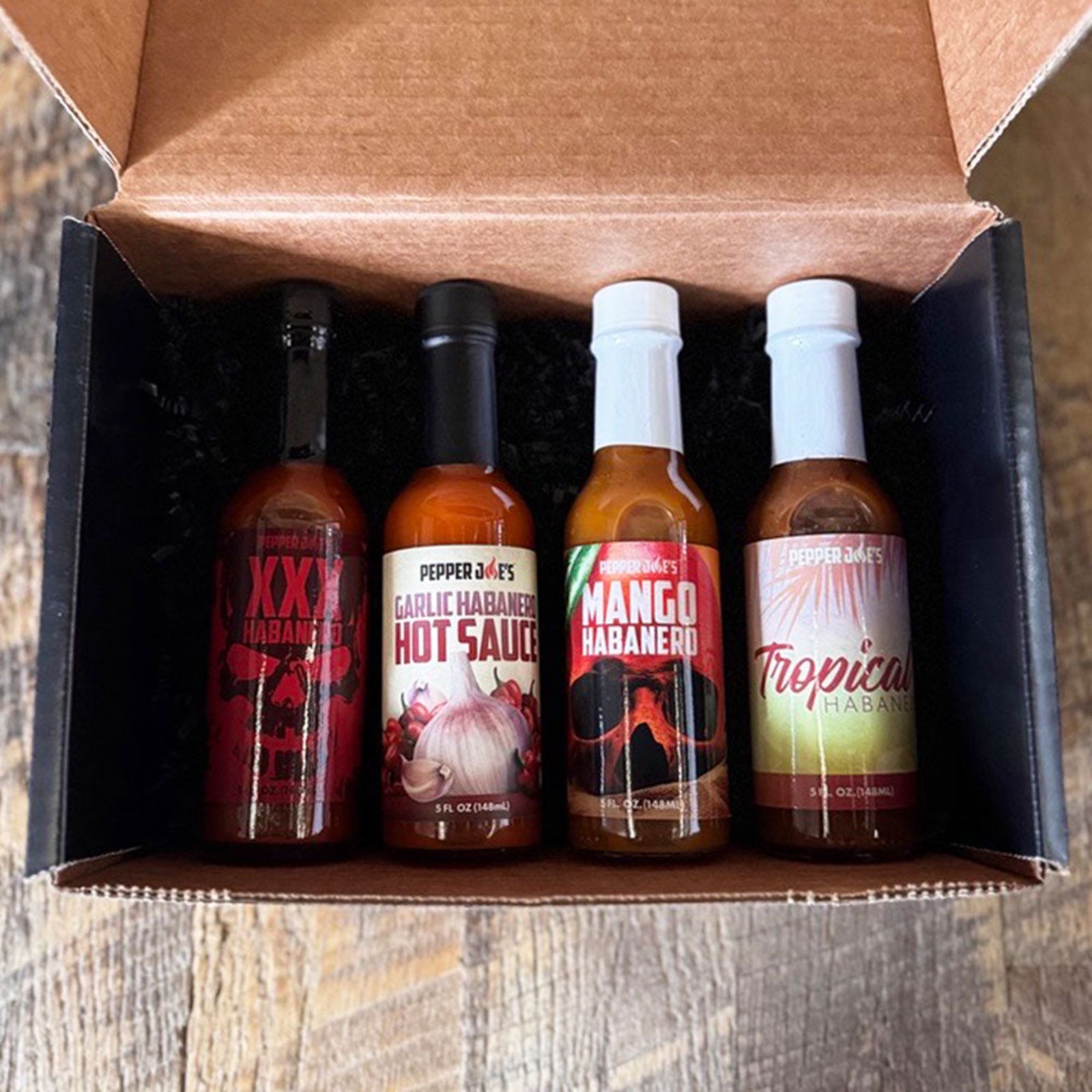 Pepper Joe's Habanero hot sauce gift box that includes one bottle each of XXX Habanero, Garlic Habanero, Mango Habanero, and Tropical Habanero inside gift box on a rustic background.