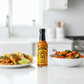 Habanero Hot Sauce bottle on a white kitchen counter beside a bowl of grilled vegetables and a plate of tacos  drizzled with spicy hot sauce.