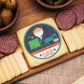 A cheese board featuring a slice of Precious Cargo Horseradish Cheddar cheese with a branded label on top, surrounded by sliced cheese, summer sausage, and olives