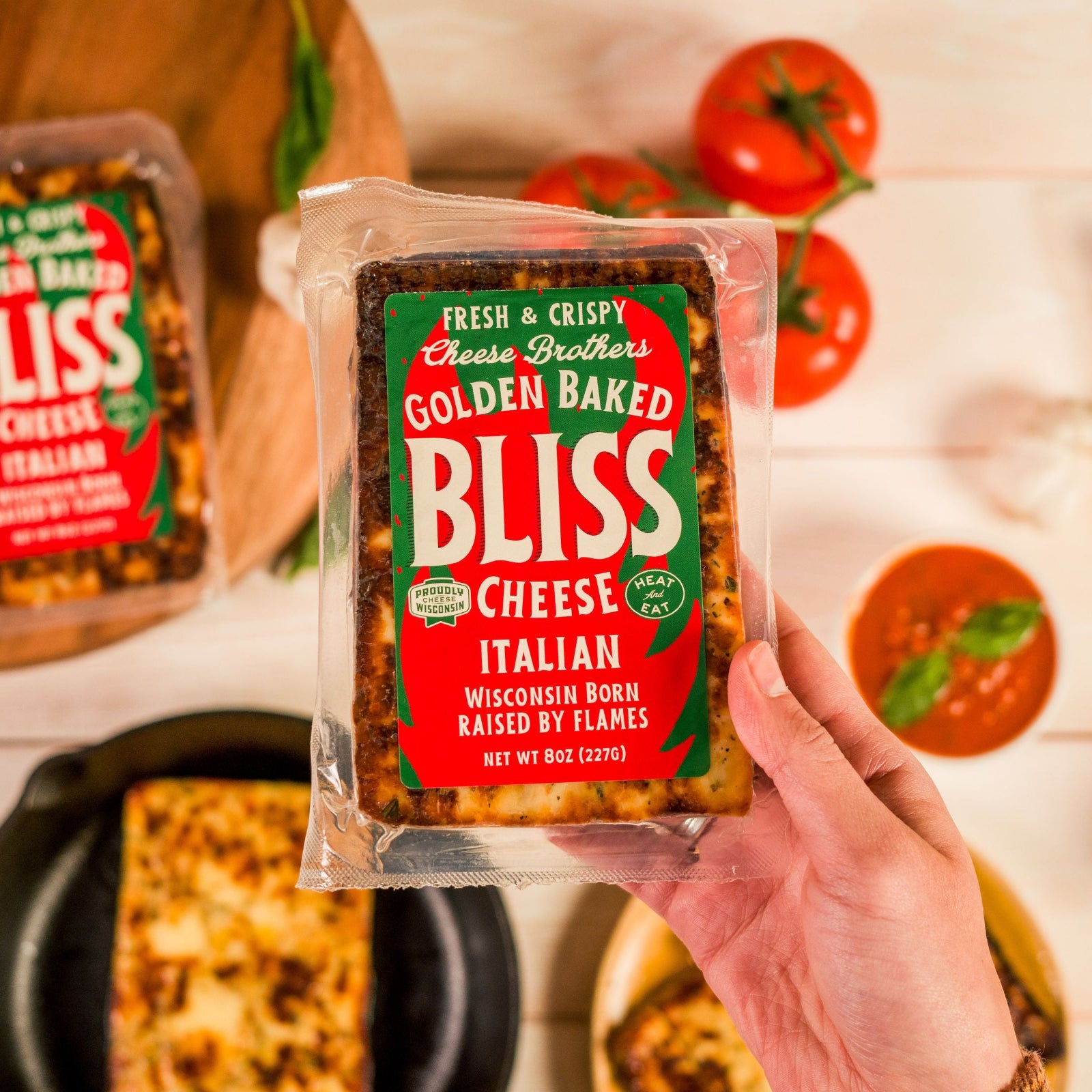 Packaged Italian Golden Baked Bliss Cheese held in front of a backdrop of tomatoes and herbs.