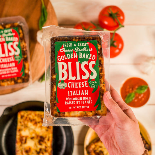 Packaged Italian Golden Baked Bliss Cheese held in front of a backdrop of tomatoes and herbs.