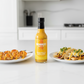 Pineapple & Habanero Hot Sauce bottle on a white kitchen counter beside a plate of grilled shrimp and a plate of tacos drizzled with hot sauce.