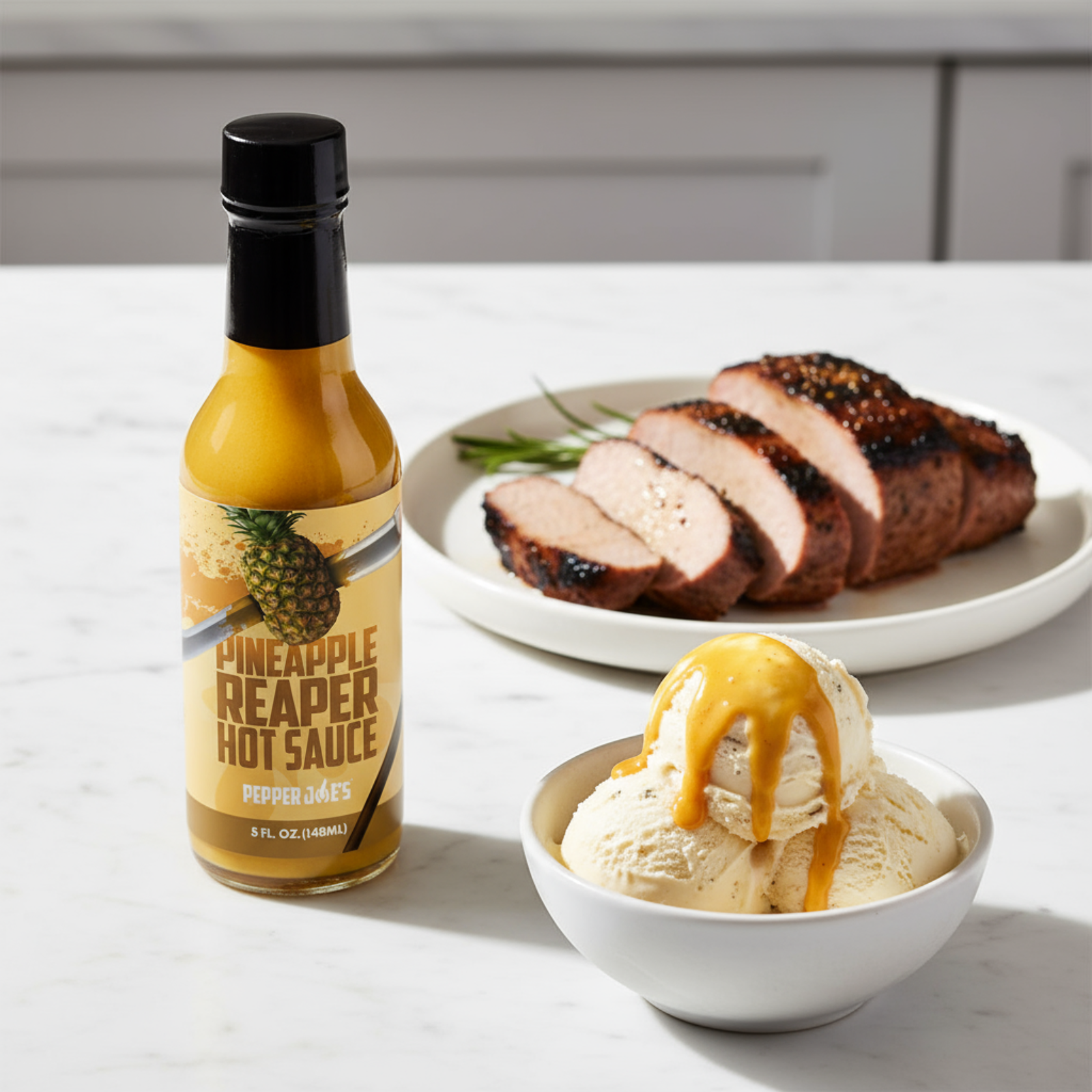 Pineapple Reaper Hot Sauce bottle on a white kitchen counter beside a bowl of ice cream and a plate of sliced pork drizzled with spicy pineapple hot sauce.
