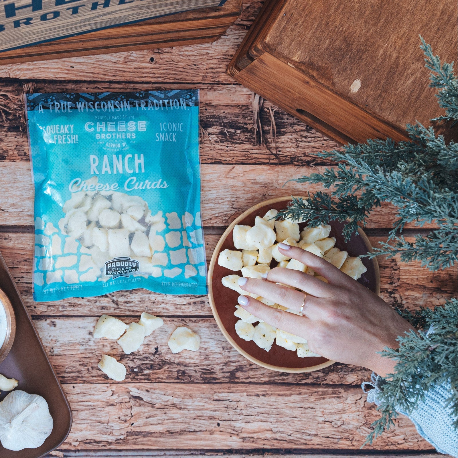 Bag of ranch cheese curds on a wooden surface with a hand reaching into bowl of curds nearby
