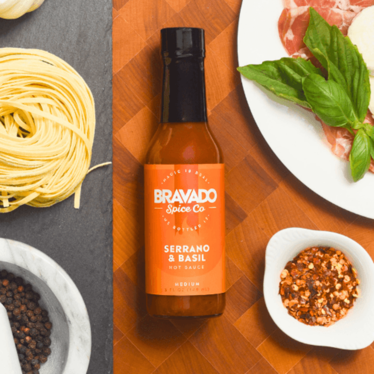 Serrano & Basil hot sauce bottle on a wooden cutting board with pasta, hot pepper flakes, and herbs.