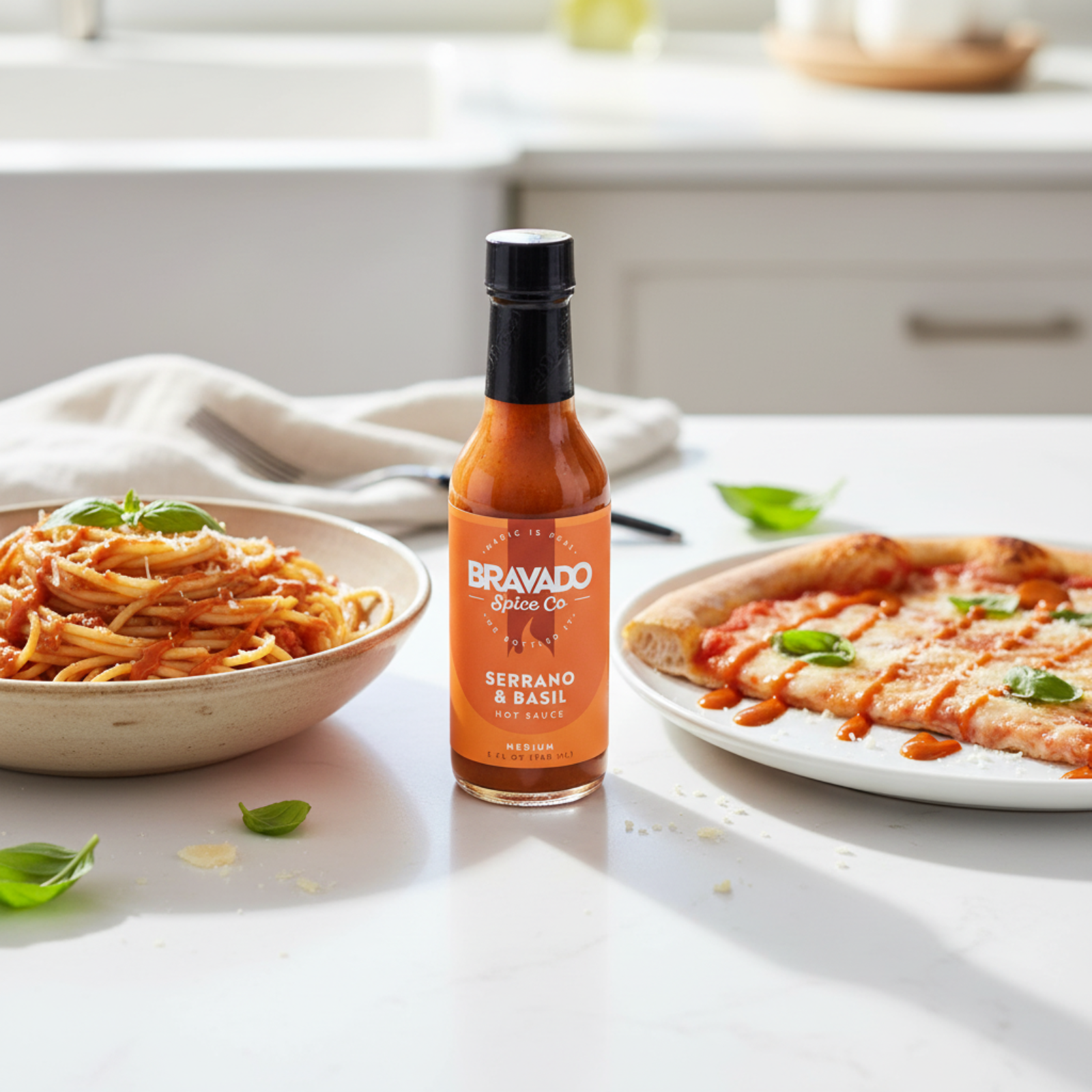 Serrano and Basil Hot Sauce bottle on a white kitchen counter beside a bowl of spaghetti and a plate of pizza drizzled with spicy blueberry hot sauce.