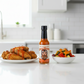 Smoked Mango Habanero Hot Sauce bottle on a white kitchen counter beside a grilled vegetables and a plate of fried chicken wings drizzled with spicy hot sauce.