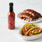 XXX Habanero Hot Sauce bottle on a white kitchen counter beside a plate of tacos and a plate of grilled pork drizzled with spicy hot sauce.